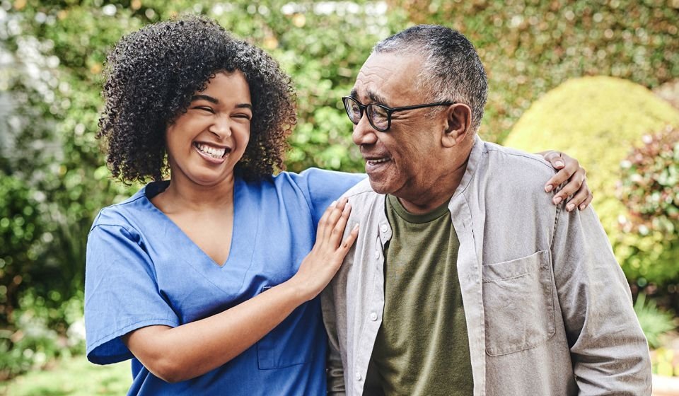 An older man happily walks with his caregiver, her arms around his shoulders.