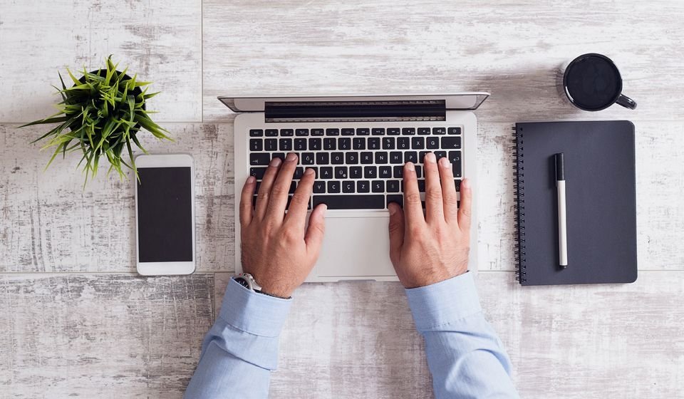 A pair of hands typing at a laptop, with a phone and notepad on either side.